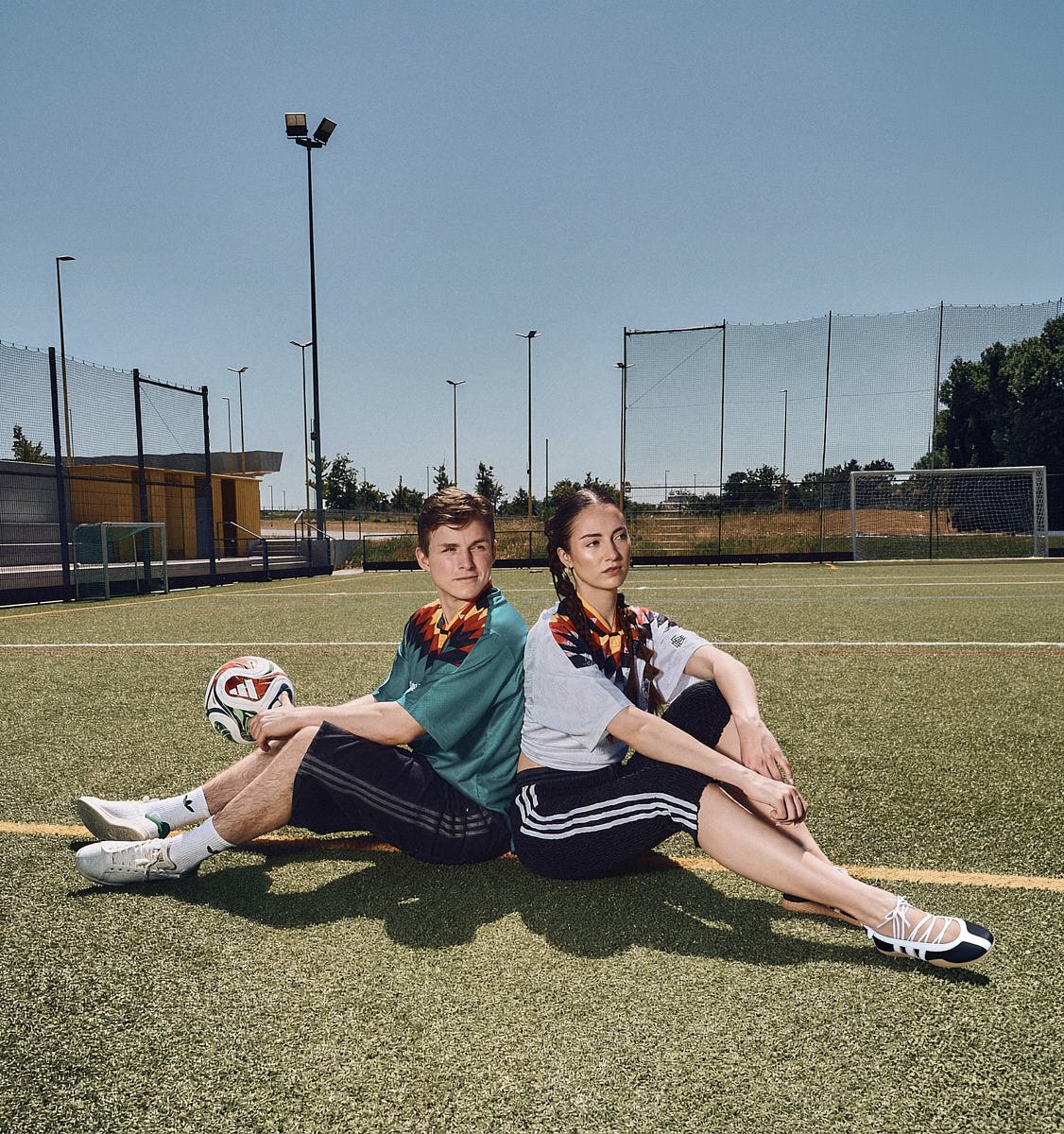 Two models sit back to back on a football pitch wearing adidas Originals Germany EQT football shirts and casual sportswear