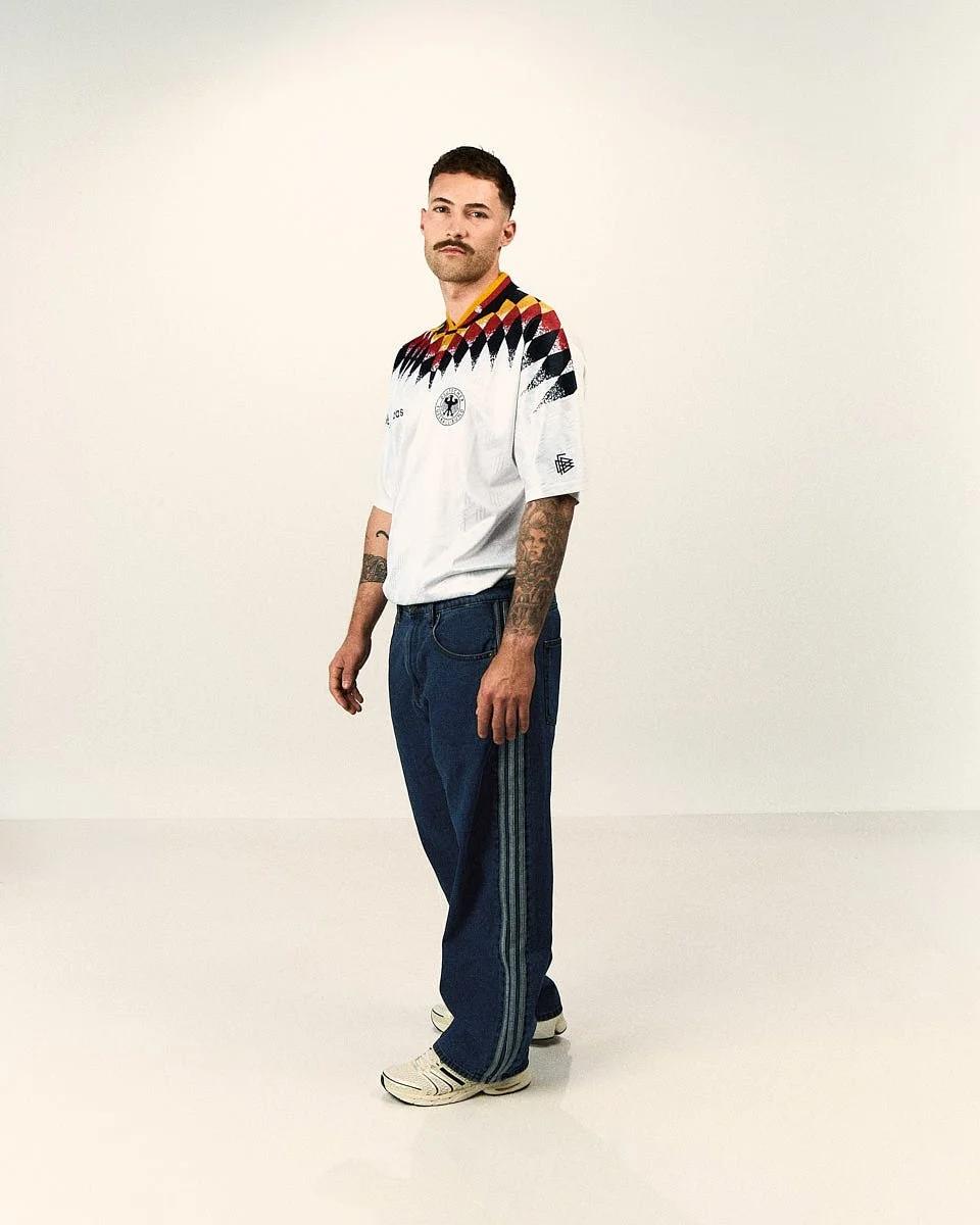 A model wears a white adidas Originals Germany EQT football shirt with wide-leg denim pants in a studio portrait