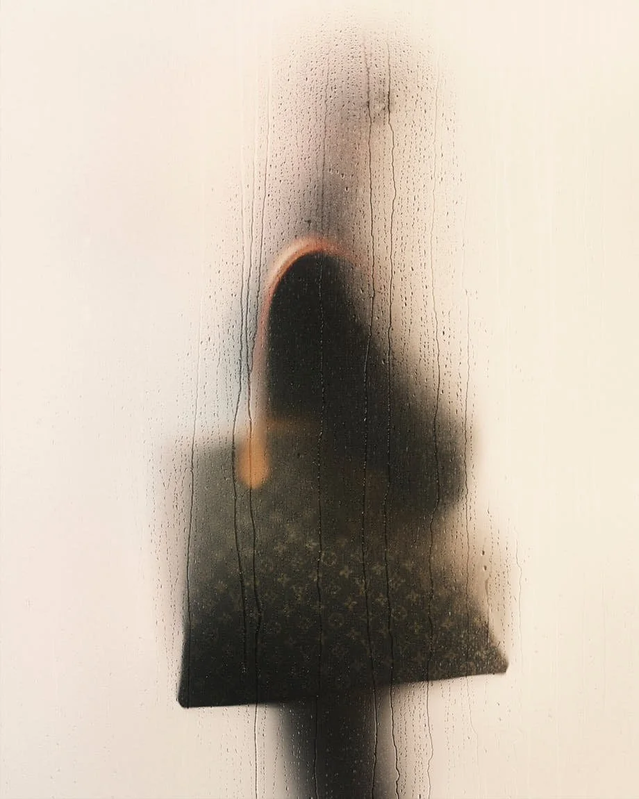 Blurred figure in dark Louis Vuitton clothing carrying a large bag behind rain-streaked glass.