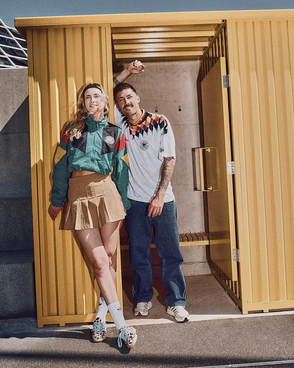 Two models wear adidas Originals Germany EQT pieces while standing in front of a yellow structure near a football pitch