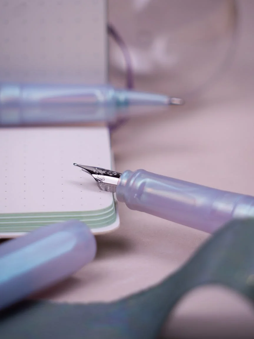 Kaweco Perkeo Iridescent Pearl fountain pens and rollerballs arranged with notebooks, packaging, and iridescent fabric on a pastel pink surface