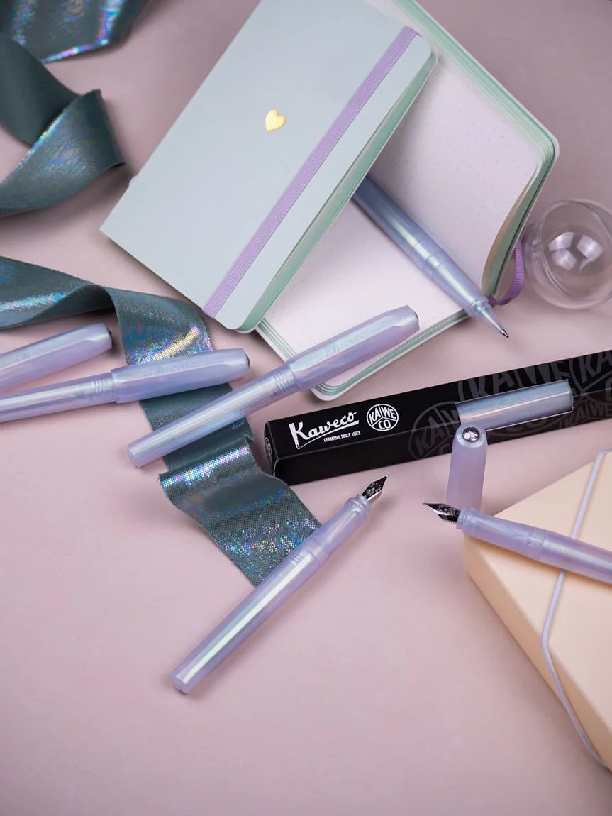 Kaweco Perkeo Iridescent Pearl fountain pens and rollerballs arranged with notebooks, packaging, and iridescent fabric on a pastel pink surface
