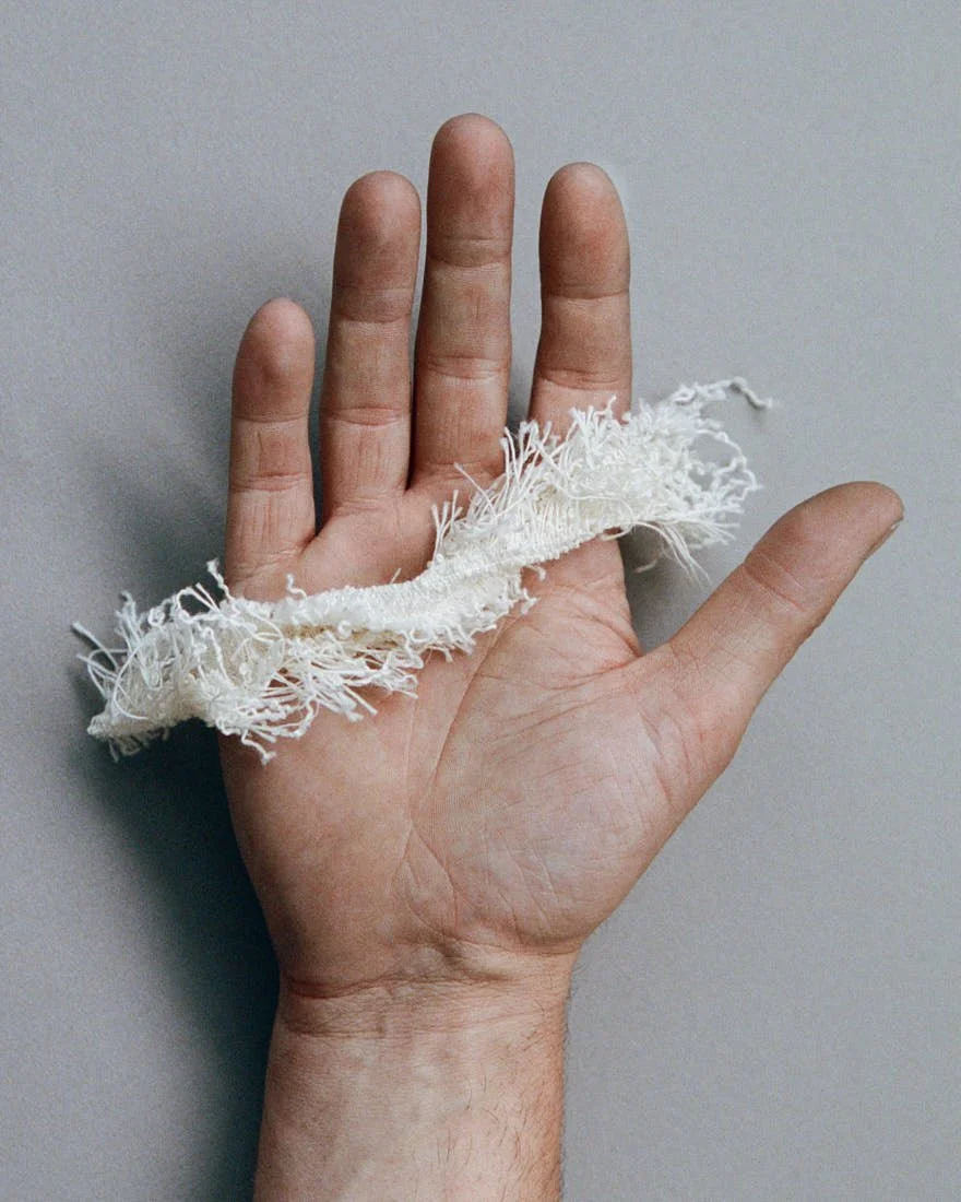 Frayed white textile sample from the Dior Men Summer 2027 collection shown against a black background and held across a hand.