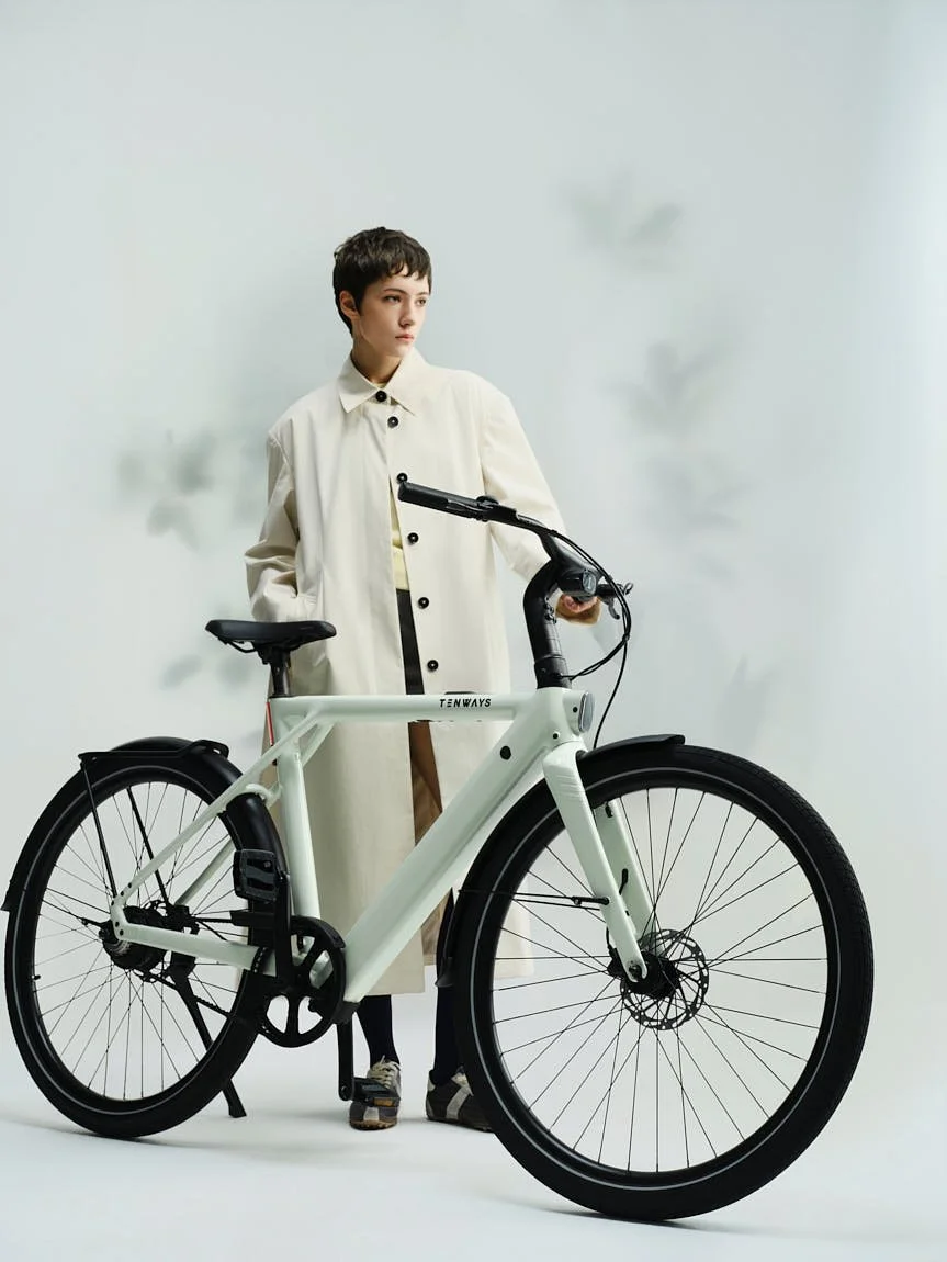 Model standing beside the pale green TENWAYS CGO 009 e-bike in a studio setting, holding the handlebar and wearing a cream coat.