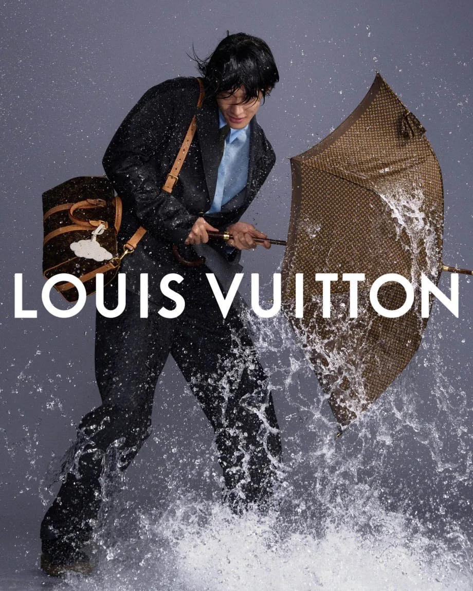 Model in dark Louis Vuitton tailoring holding a brown umbrella amid splashing water.