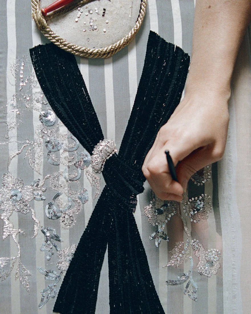 Close-up of Dior Men Summer 2027 embroidery work, showing silver beadwork, a black bow detail and hand-applied embellishment on striped fabric