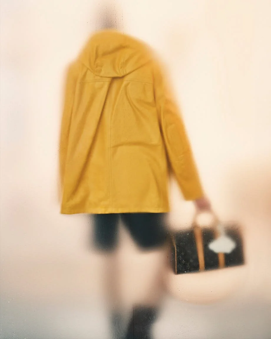 Rear view of a yellow Louis Vuitton hooded jacket and dark Monogram bag through rain-streaked glass.