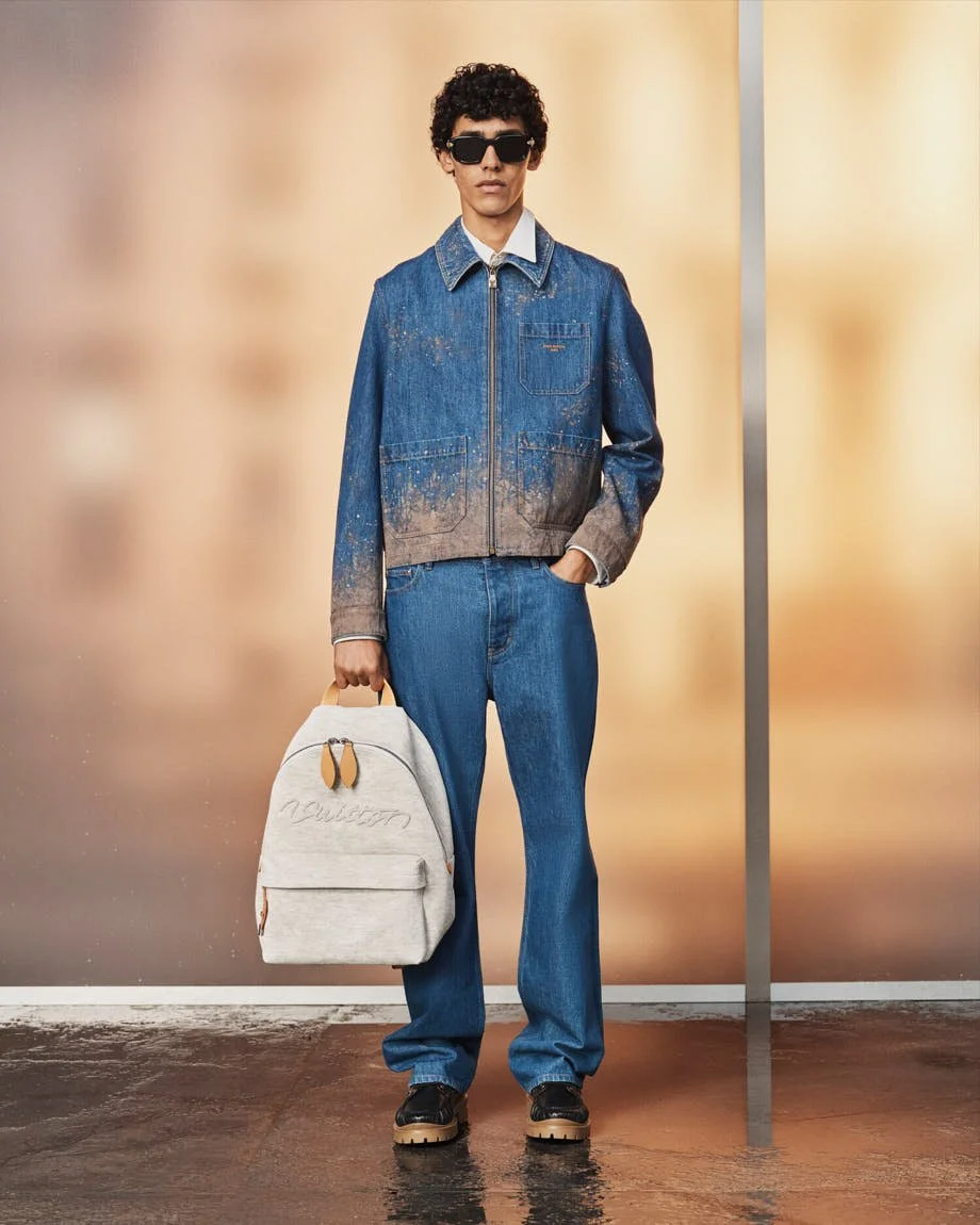 Model wearing a mud-effect Louis Vuitton denim jacket with blue jeans and carrying a pale grey backpack.