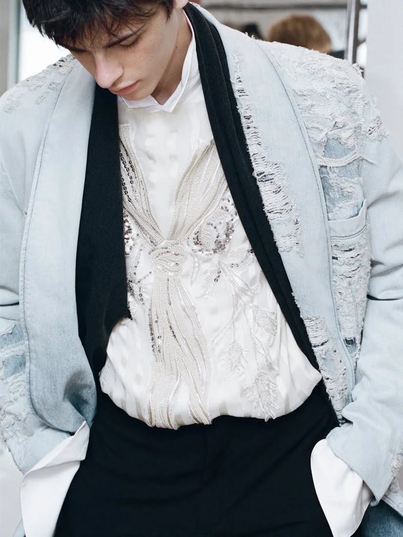 Close-up of a model backstage at Dior Men Summer 2027 wearing a pale denim jacket with frayed detailing over an embroidered white silk shirt and black scarf for LE MILE Magazine