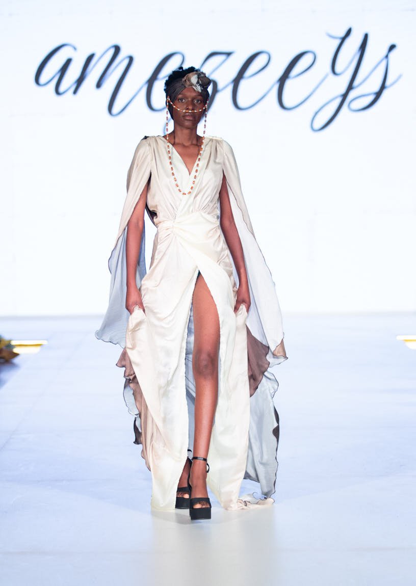 Model walking the runway in a flowing white dress with a high slit and draped detailing at Zimbabwe Fashion Week.