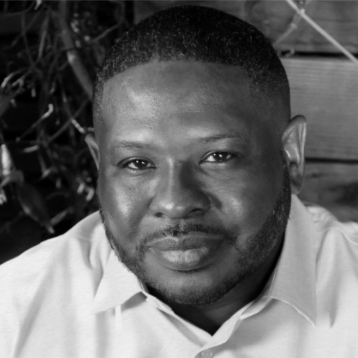 Close-up of a black man with short hair, a beard, and wearing a collared shirt, looking at the camera.
