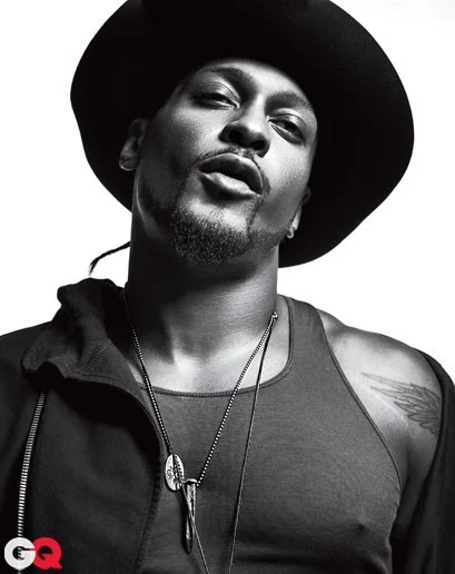 GQ cover of D'Angelo, a black man wearing a wide-brim hat, a tank top, and a necklace, with a serious expression and a tattoo on their shoulder, in black and white.