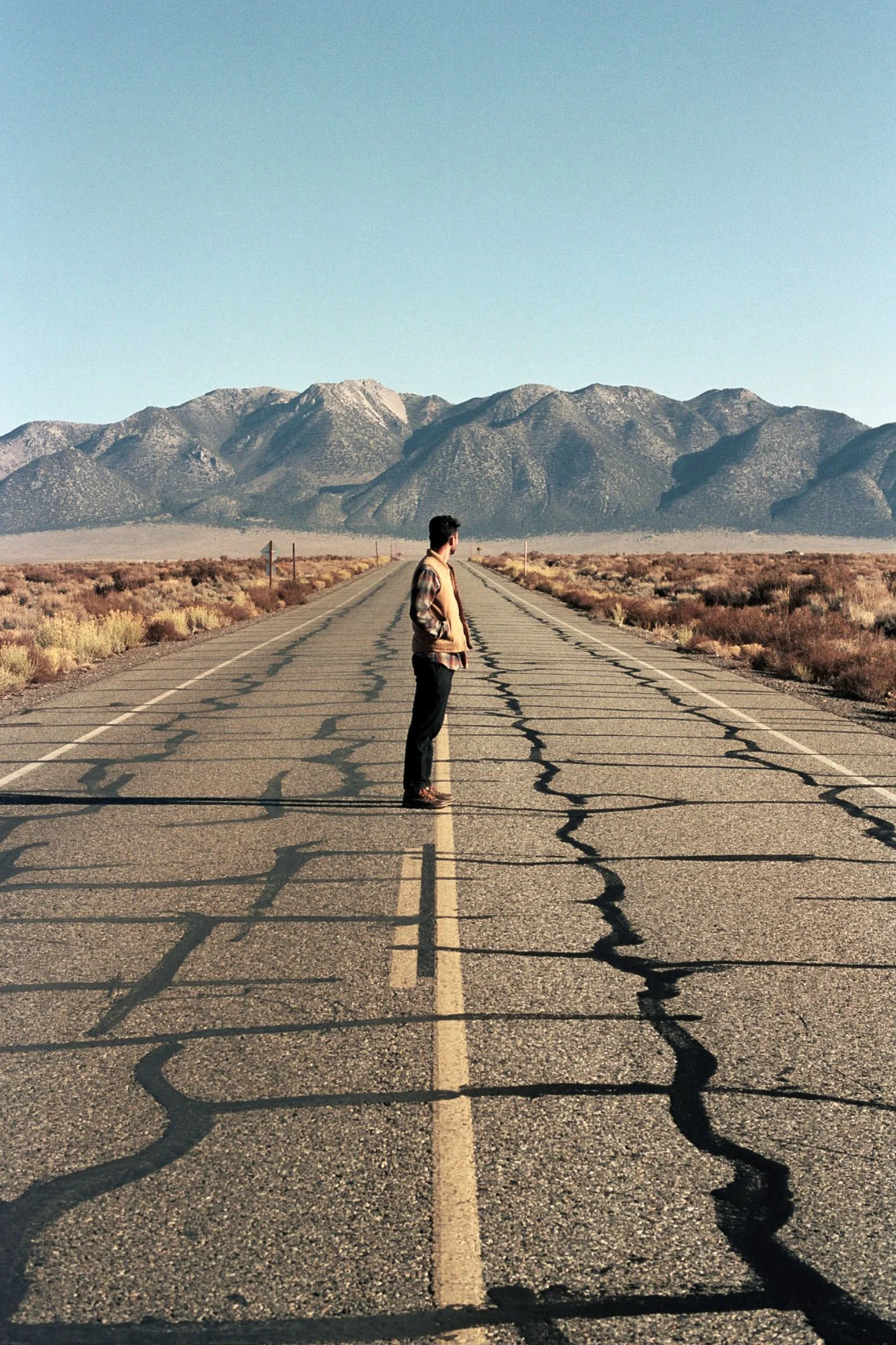 A person standing alone on a cracked desert highway that stretches into the horizon with mountains in the background and a clear blue sky travel imagery photographed by Los Angeles based, Maui born photographer and director Bailey Rebecca Roberts