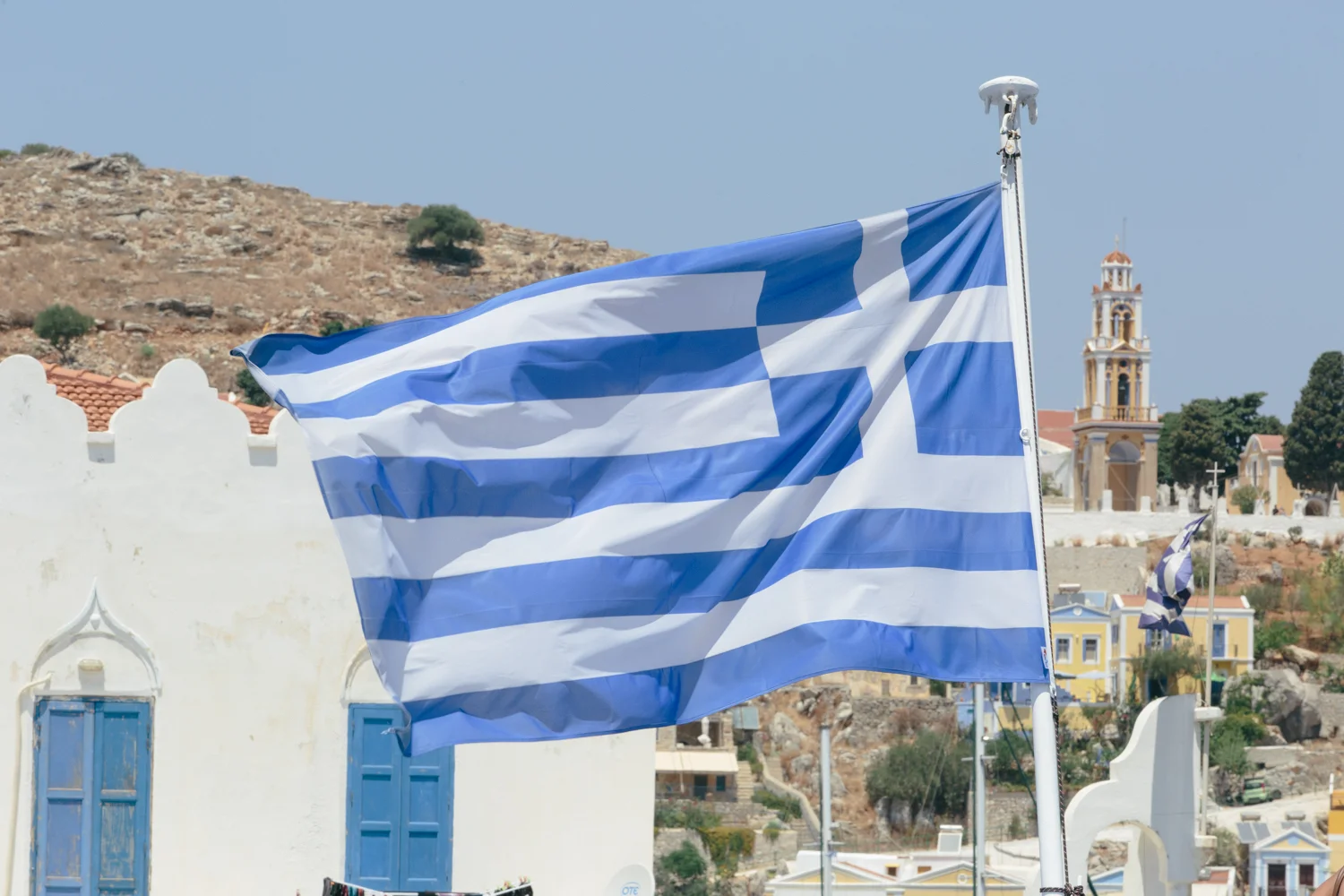 Bandeira da Grécia ondulando ao vento, com casas de paredes brancas e telhados vermelhos ao fundo, e uma colina árida ao longe.