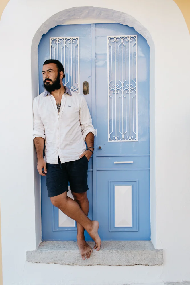 Homem de cabelo escuro, barba, vestindo camisa branca de mangas roll-up e bermuda azul, apoiado na parede ao lado de uma porta azul com grades brancas na casa de paredes brancas e moldura arredondada.