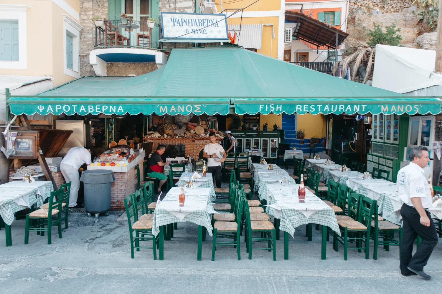 Restaurante com mesas cobertas por toalhas de xadrez em cima da rua, com uma grande cobertura verde, ambiente ao ar livre, garçom atendendo e decoração típica grega.