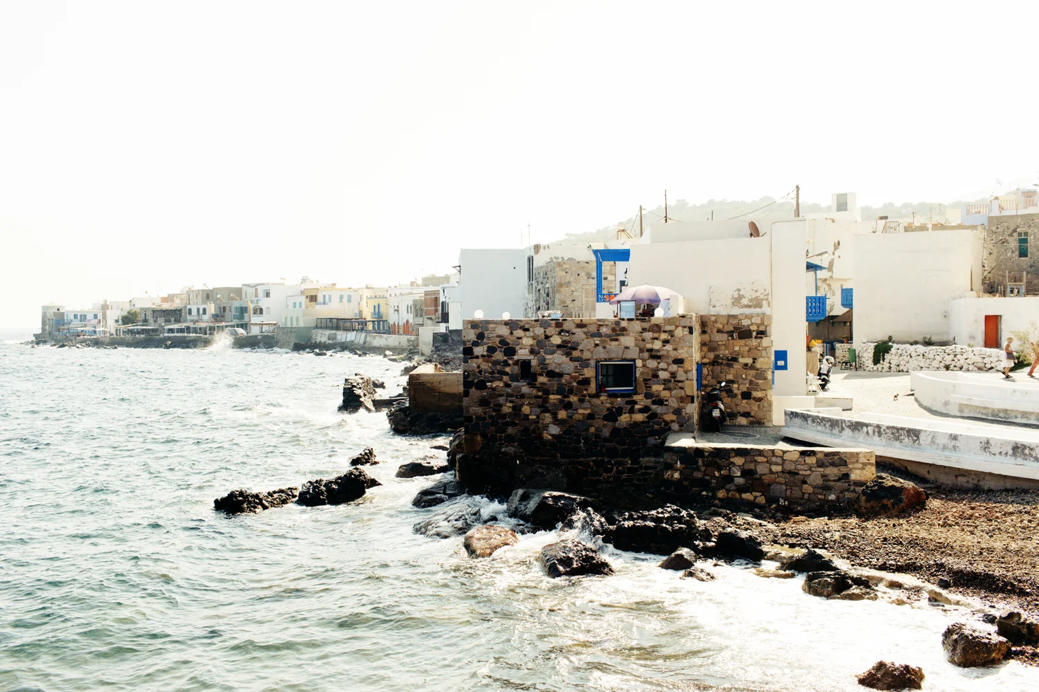 Casas brancas ao longo de uma costa, com o mar na frente e rochas ao redor, sob céu claro.