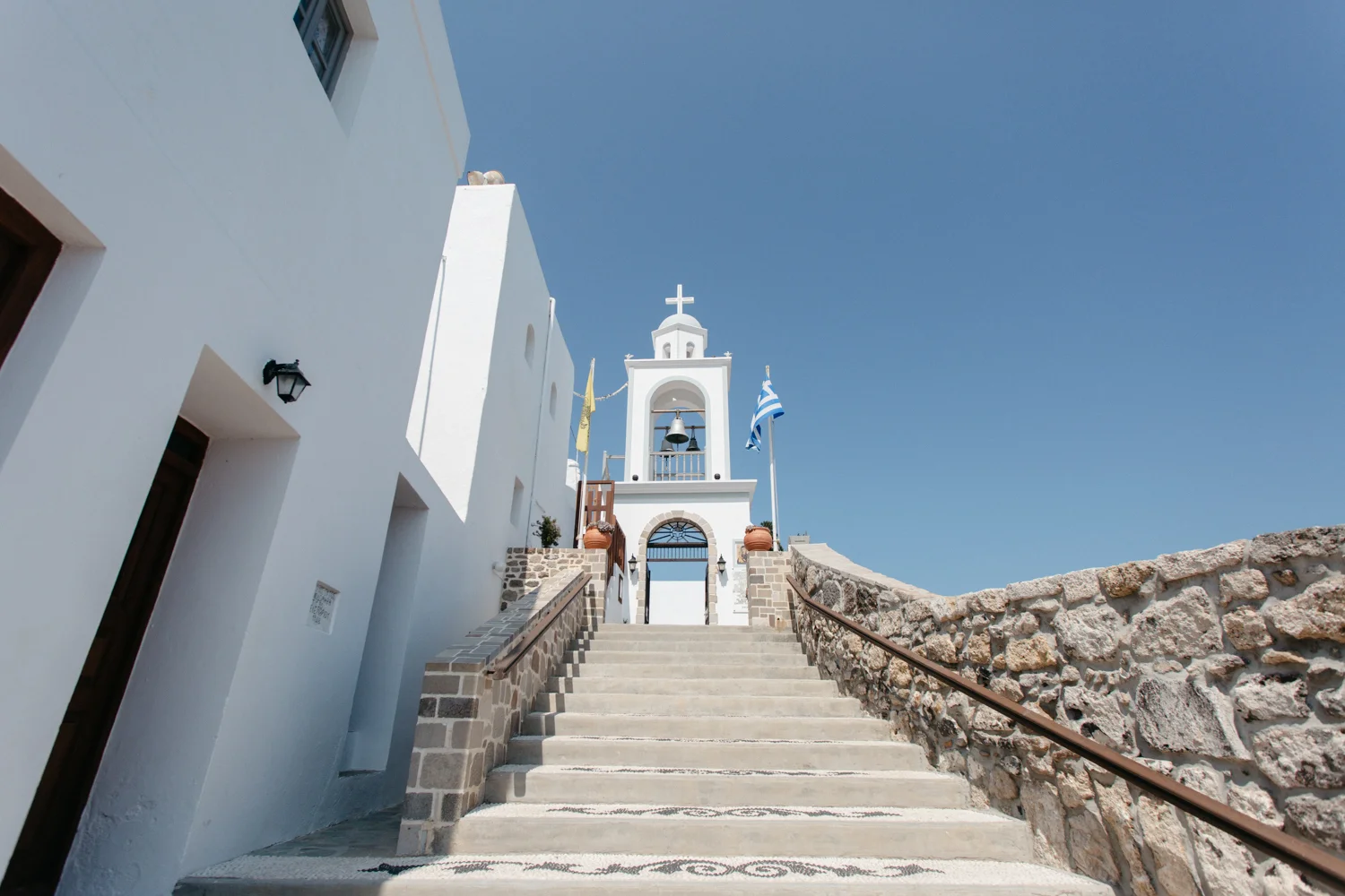 Escadaria de pedras levando a uma capela branca com cruz no topo, bandeiras gregas e céu azul ao fundo