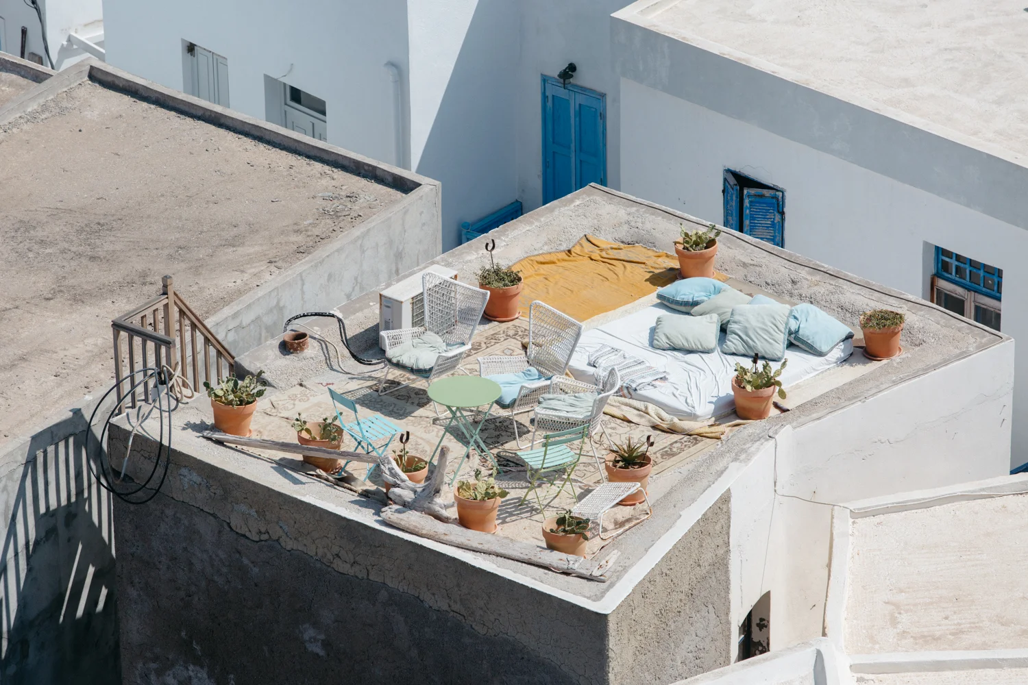 Terraço com pufes, cadeiras de metal, almofadas, uma manta amarela, várias plantas em vasos de barro, uma roupa de cama e uma toalha cobrindo parte do piso, em um prédio com paredes brancas e janelas de madeira azul.