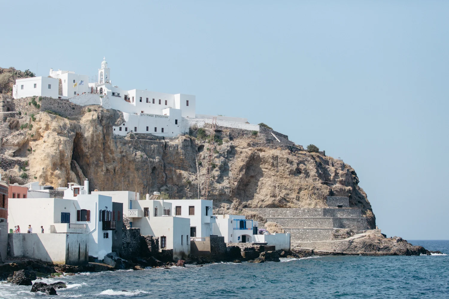 Casas brancas tradicionais de Santorini, na Grécia, situadas em penhascos à beira-mar com o oceano em primeiro plano e céu claro ao fundo.
