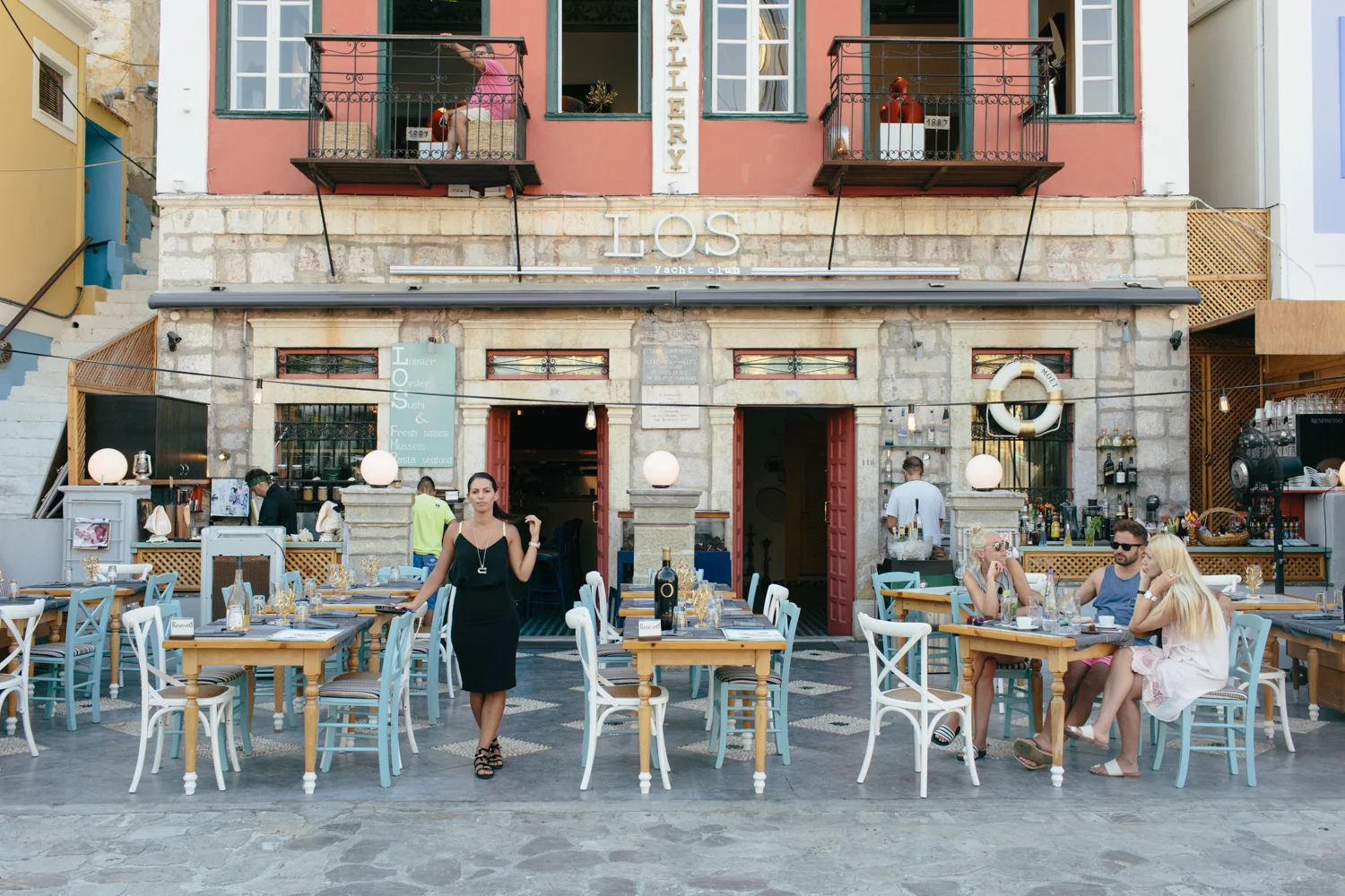 Restaurante ao ar livre com mesas e cadeiras, ambiente decorado com luzes e decoração marítima, pessoas jantando e uma mulher de vestido preto na frente, fachada de edifício antigo com sinalização de bar e restaurante.
