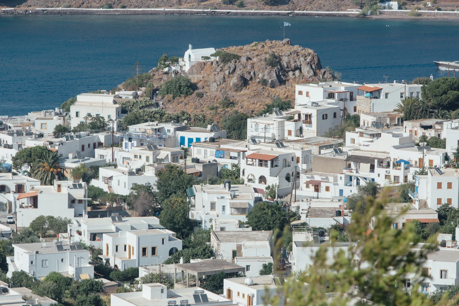 Cidade de casas brancas ao lado de uma água azul, com uma capela branca no topo de uma colina rochosa.