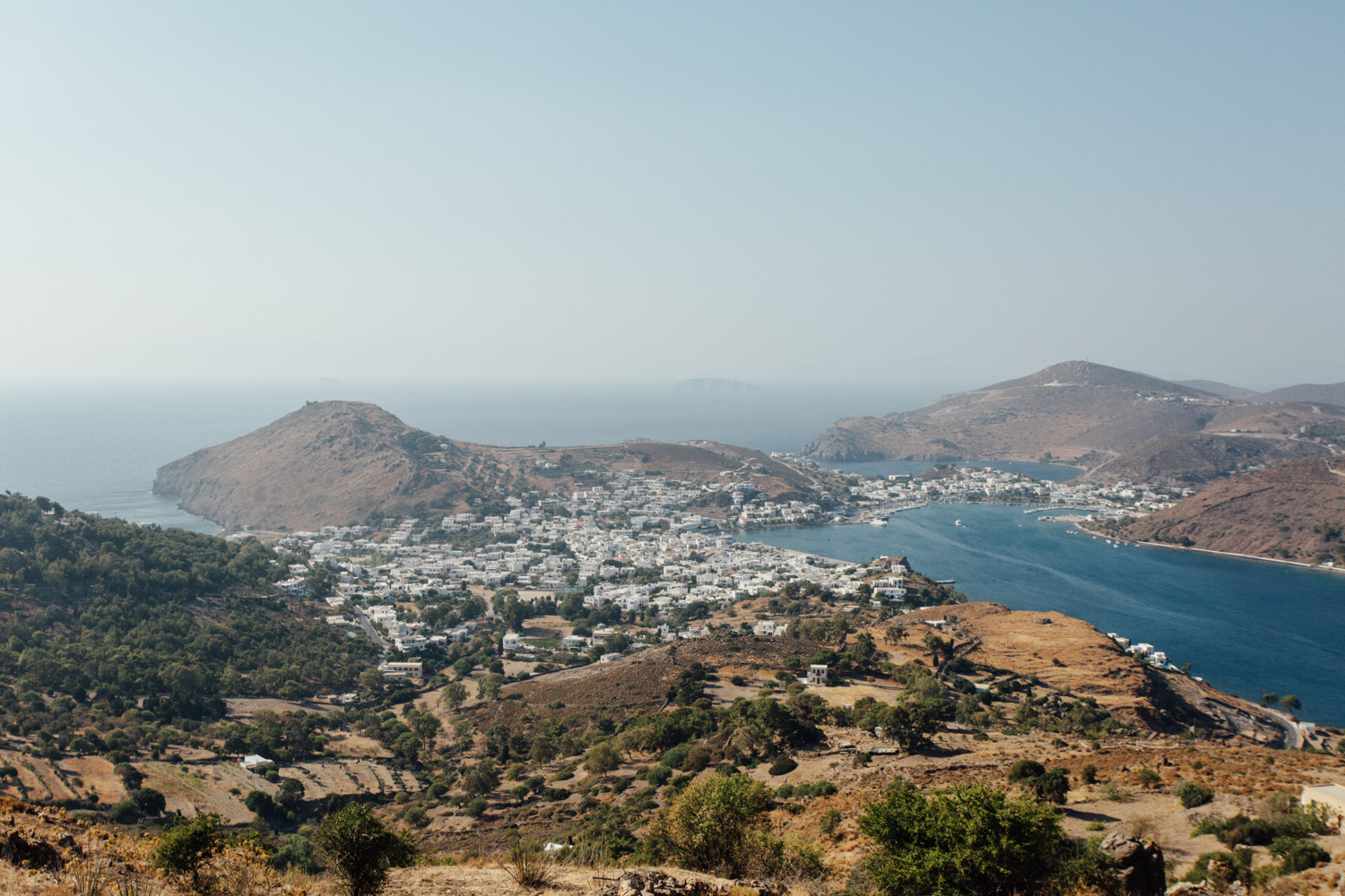 Paisagem de uma ilha com mar, montanhas áridas e vilarejo com casas brancas, vista de uma região montanhosa rural.