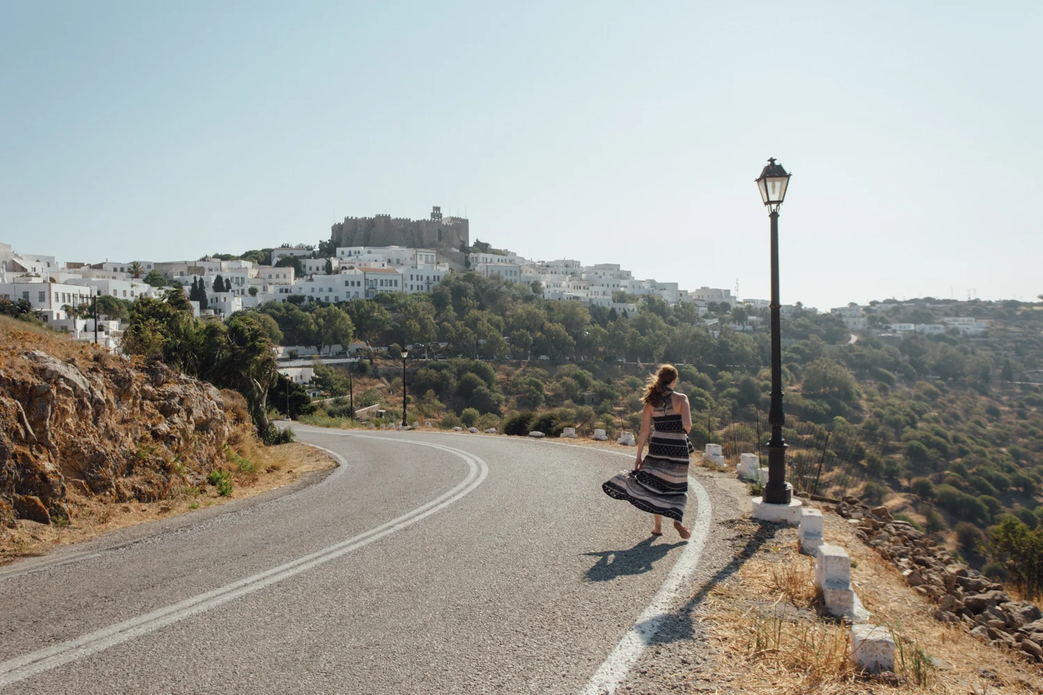 Mulher caminhando na rua de uma cidade com casas brancas em uma colina, ao entardecer, com um castelo ao fundo e uma lâmpada de rua ao lado