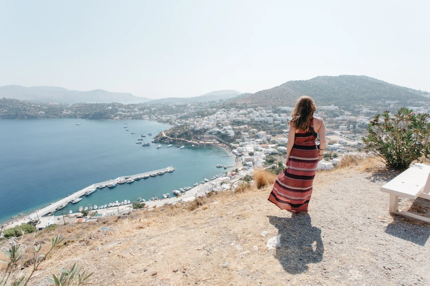 Mulher de cabelo castanho vestindo vestido vermelho e preto, de costas, olhando para uma vista de cidade e mar ao longe, em uma ilhota, com montanhas ao fundo, sentado em banco branco na encosta seca.