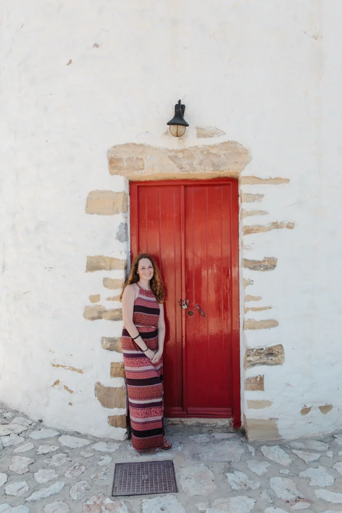 Mulher de cabelo castanho claro, vestindo vestido longo com padrão listrado em tons de vermelho, preto e branco, posando na frente de uma porta vermelha com parede branca de pedra, sob uma luminária preta na parede.