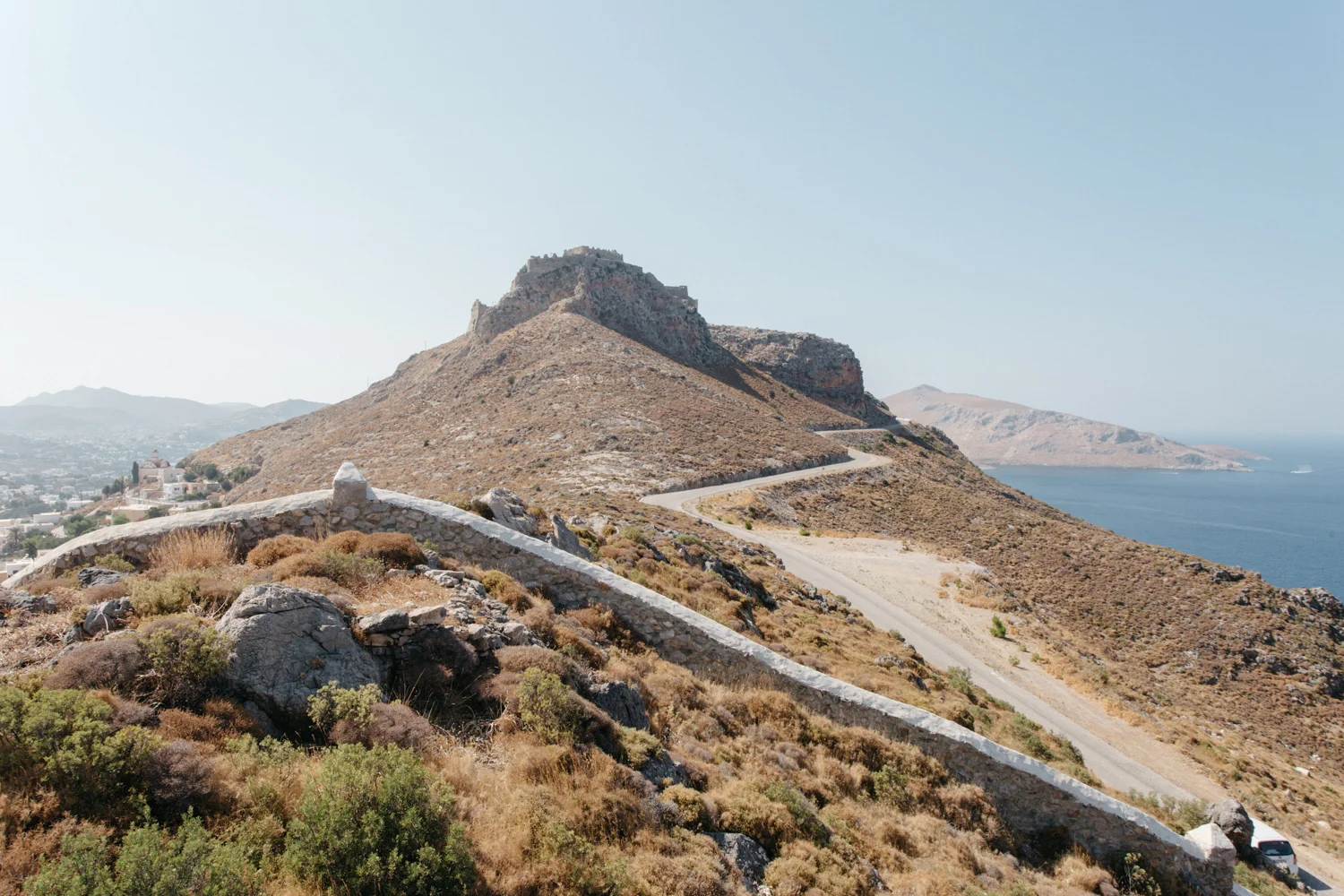 Montanha com estrutura antiga no topo, estrada serpenteando ao redor, vegetação seca no primeiro plano, vista para o mar ao fundo