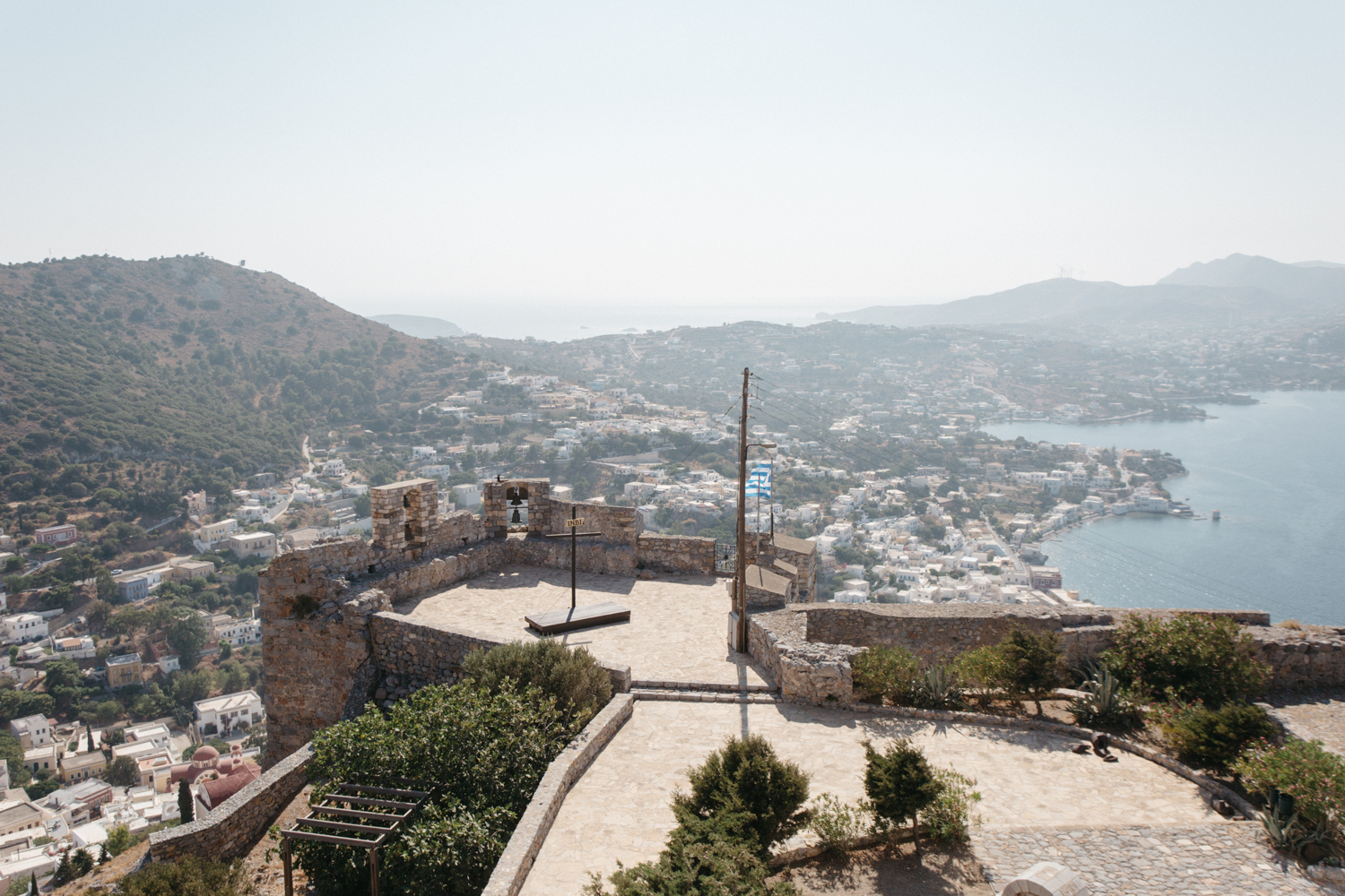 Vista de uma cidade à beira de um mar, vista de uma fortaleza antiga com pedras e vegetação, com uma bandeira da Grécia hasteada.