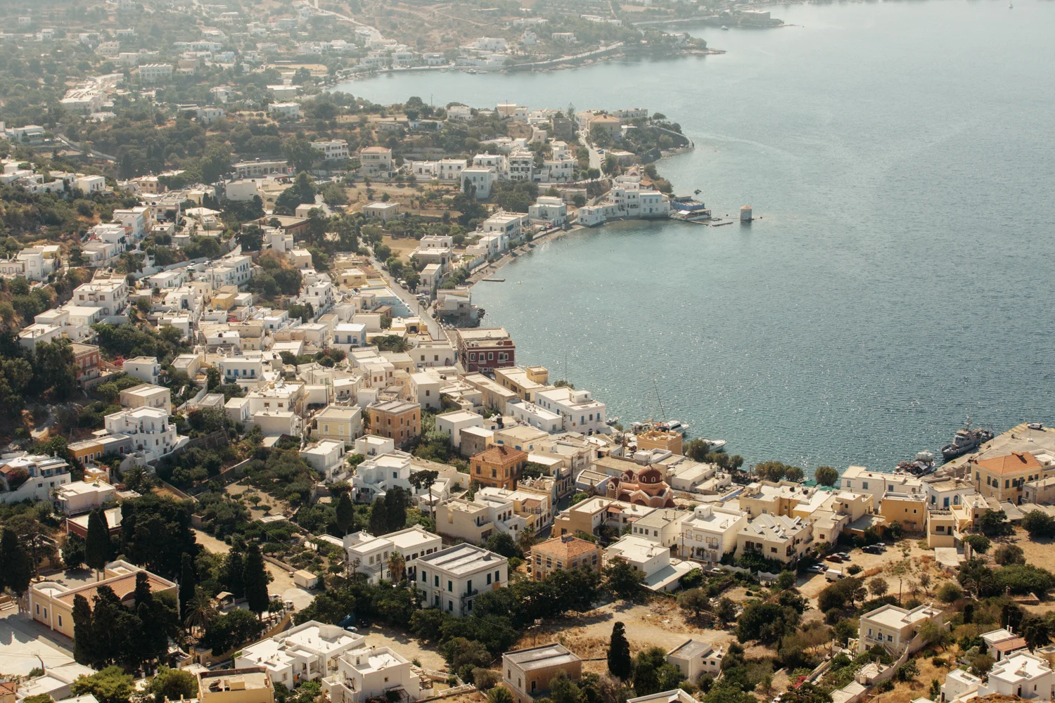 Vista aérea de uma cidade costeira com casas brancas, em um terreno levemente acidentado ao lado de uma baía. O mar ao lado apresenta barcos e pequenas embarcações. O cenário inclui vegetação dispersa e ruas estreitas.