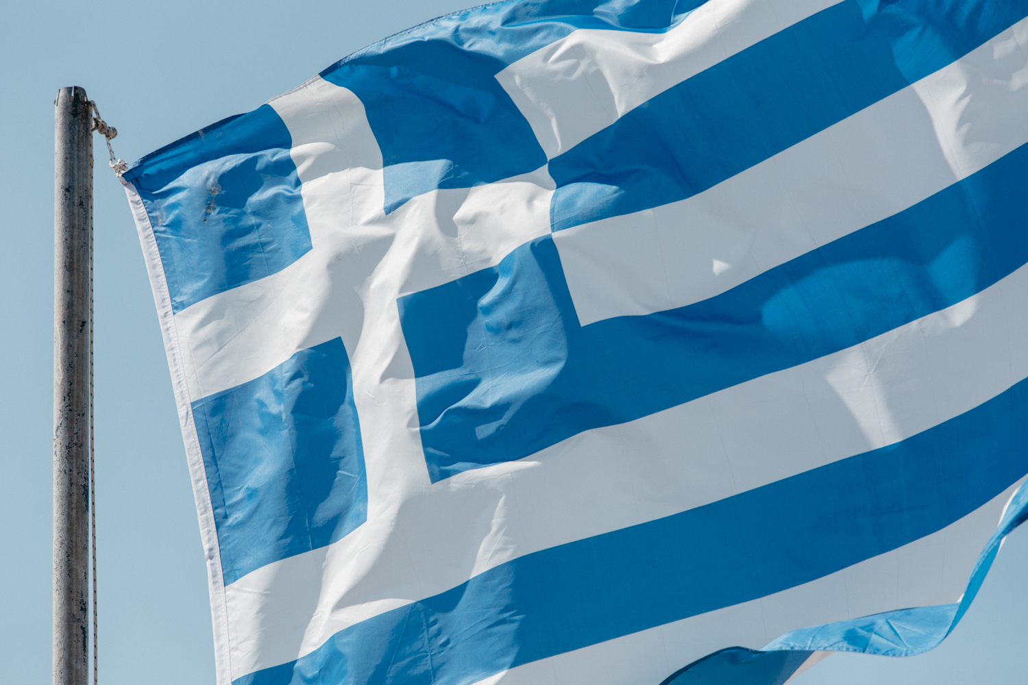 Bandeira da Grécia com listras azuis e brancas e uma cruz branca no cantinho azul, tremulando ao vento.