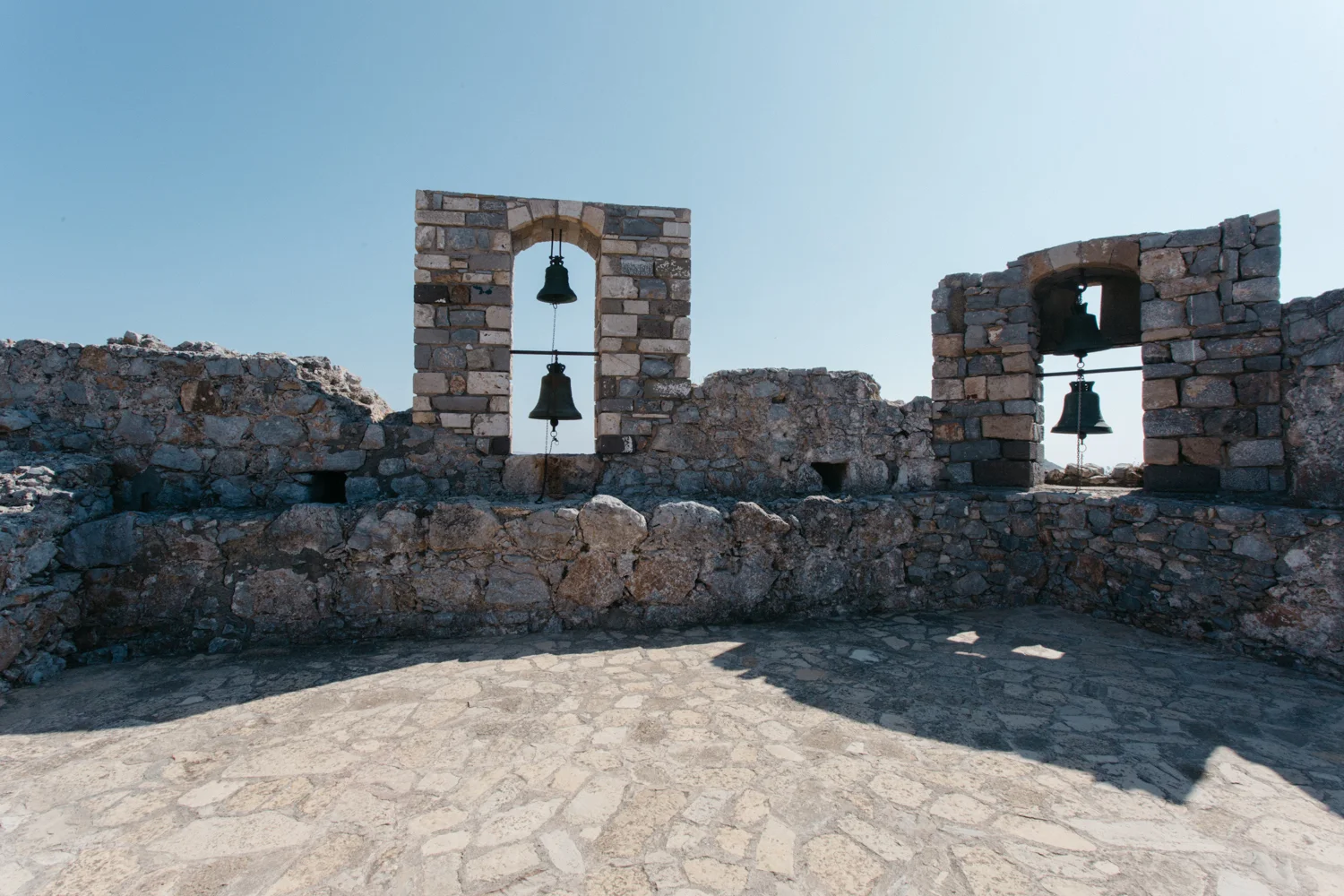 Ruínas de uma construção antiga de pedra, com duas janelas de arcos de pedra, cada uma contendo um sino de metal, sob um céu claro.