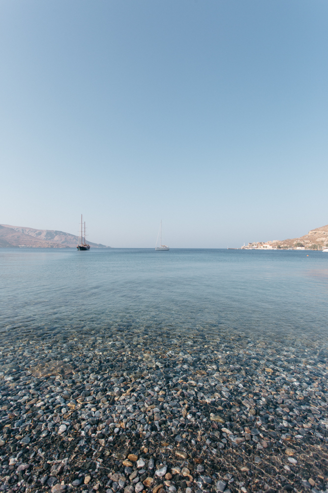 Cena de uma praia com água transparente, pequenas pedras na0or, e três veleiros ao fundo, com colinas ao redor.