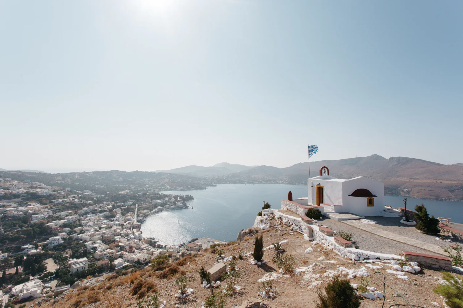 Vista de uma ilha com uma capela branca ao lado de um mar calmo, com uma bandeira da Grécia ao vento. Ao fundo, uma cidade e montanhas sob um céu claro e ensolarado.