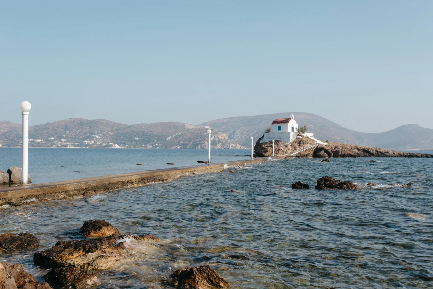 Caminho de pedra que leva a uma pequena capela branca com telhado vermelho, localizada na ilha de uma costa rochosa, com mar ao redor e montanhas ao fundo