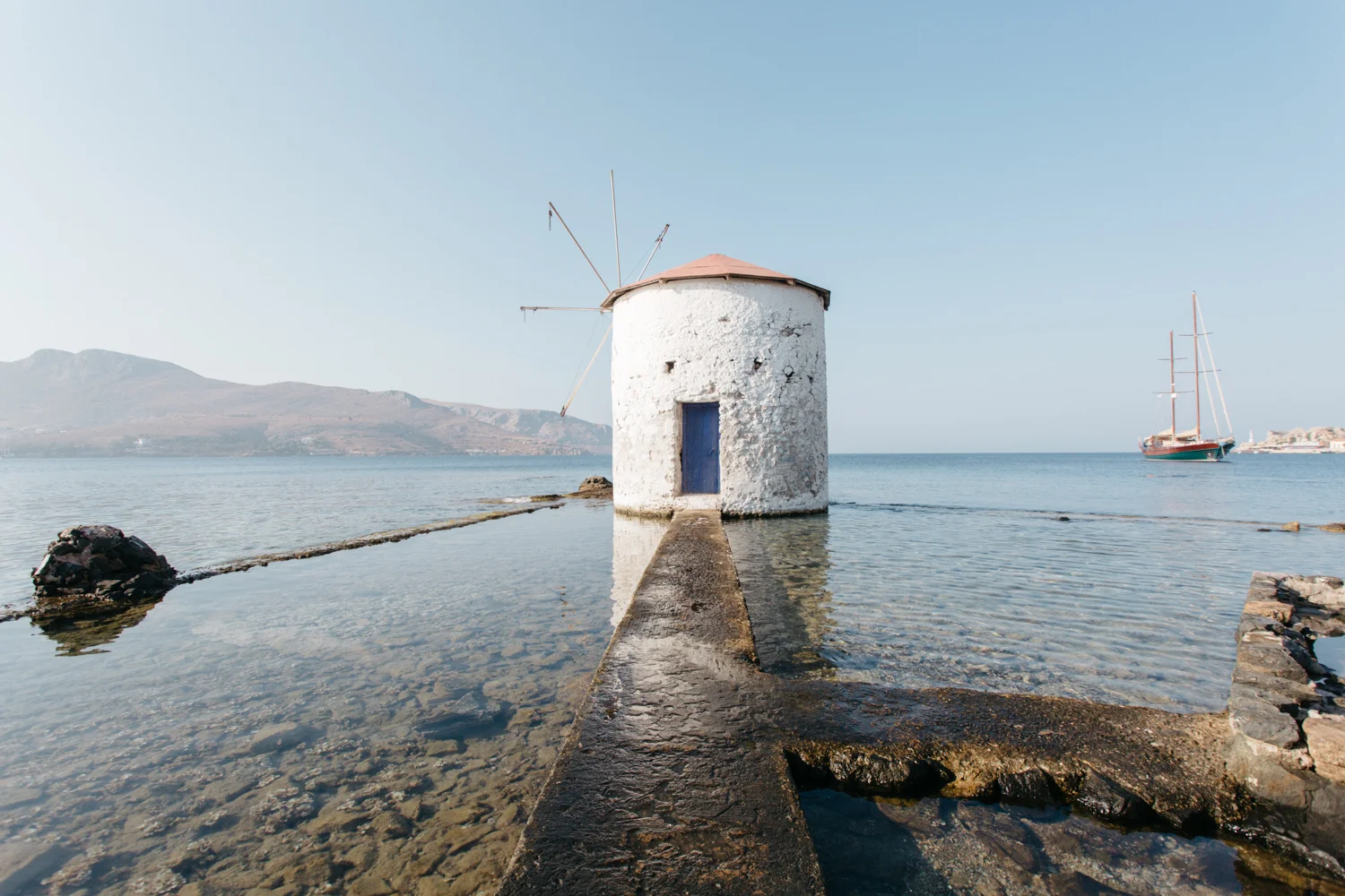 Moinho de vento branco ao lado do mar com barco de vela ao fundo.