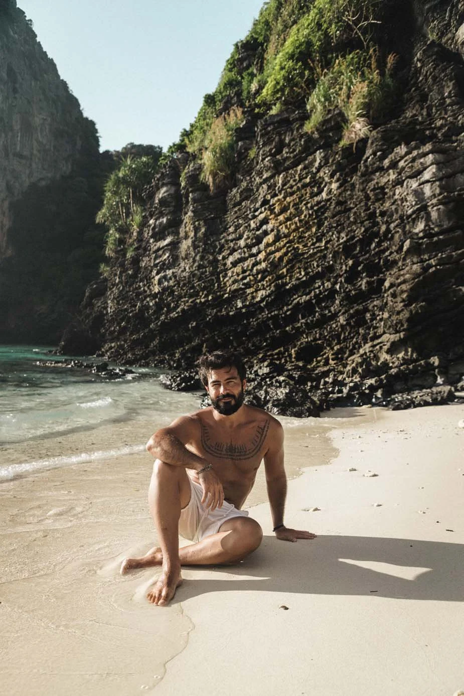 Um homem sentado na praia, sem camisola, a desfrutar do sol e da vista para o oceano.