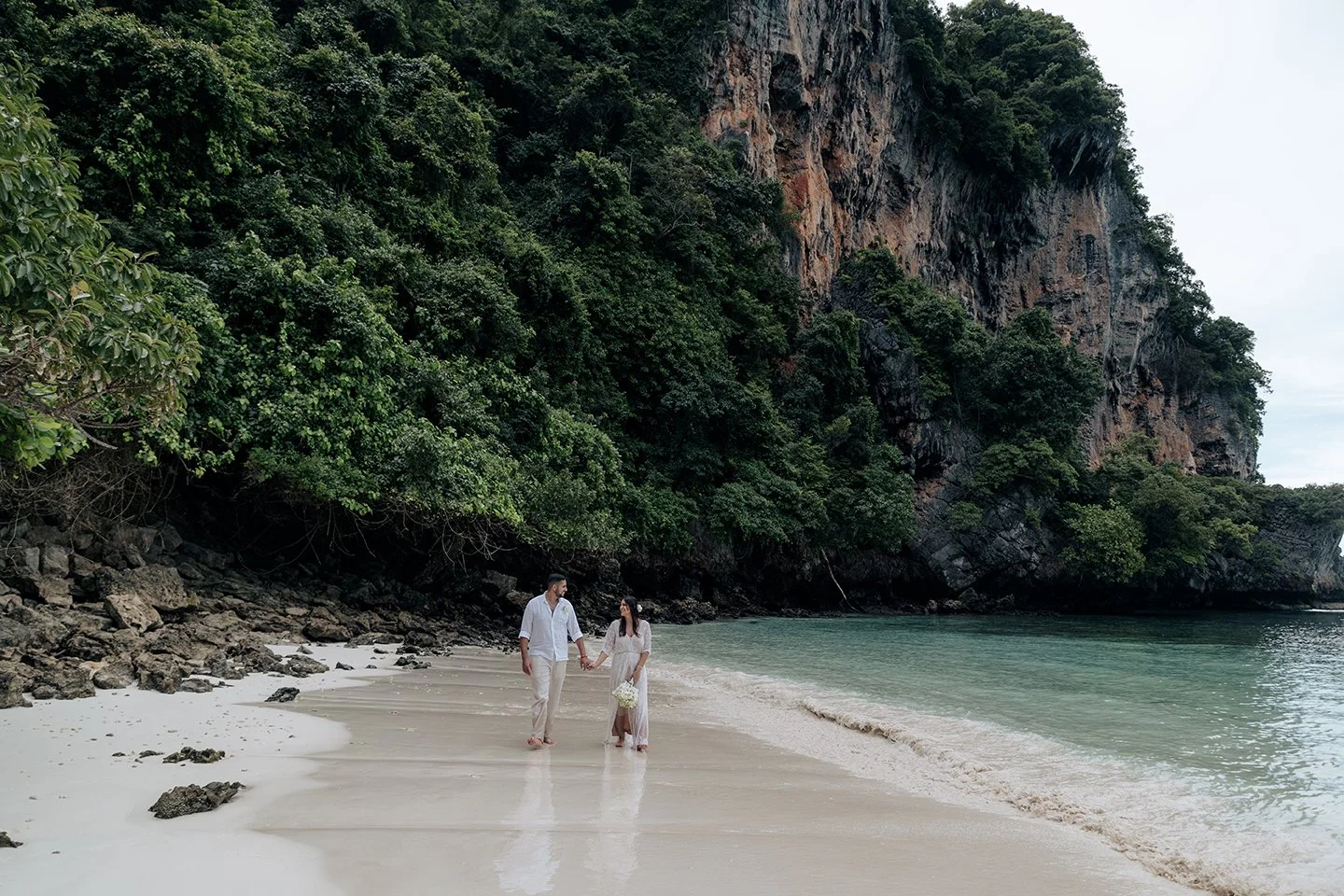 Um casal caminha pela praia, com um imponente penhasco como pano de fundo.