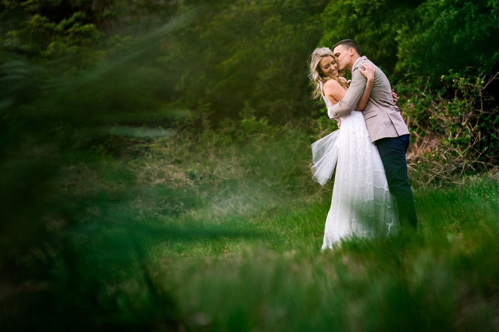 Bride smiles as groom hugs her in green forest backdrop