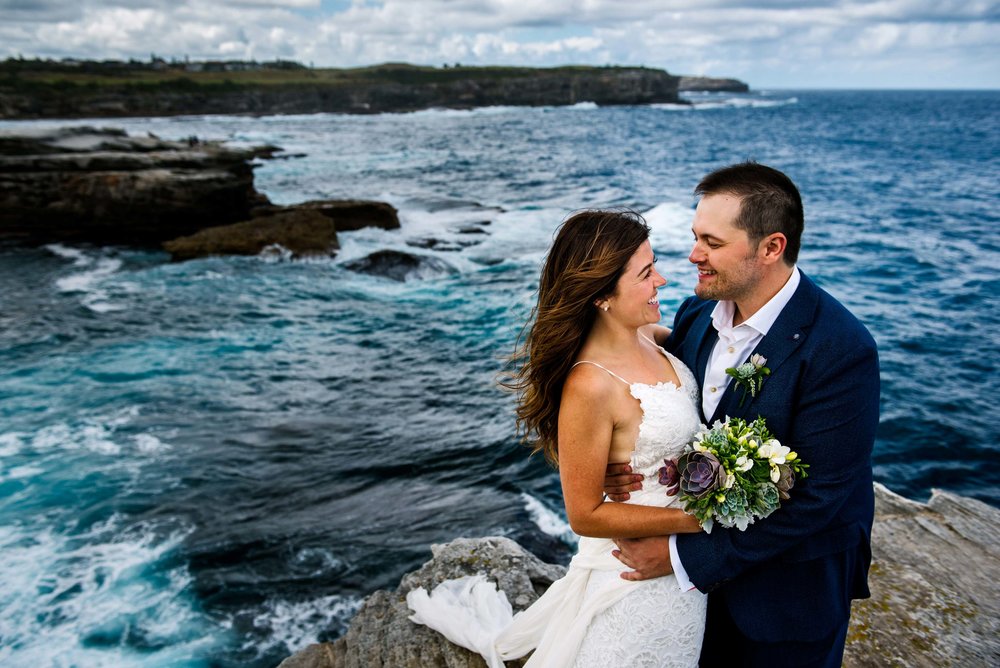 Bride and groom smile at eachother with views of Little Bay and the ocean in the background