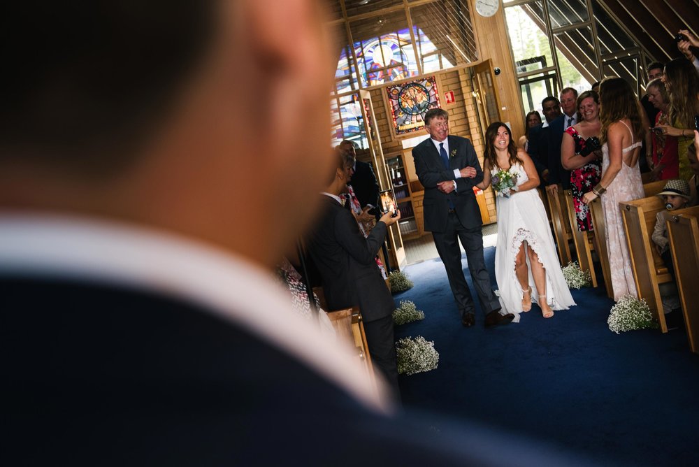 Bride and father of the bride walking down aisle with groom looking on at chapel ceremony, Little Bay