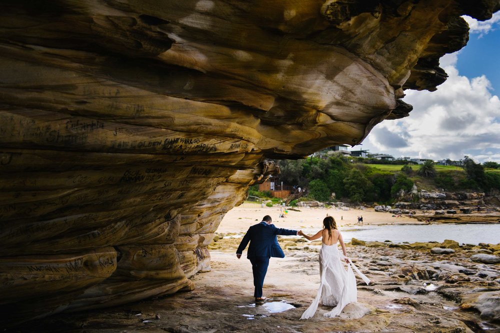 Newlyweds walk under rock outcrop along the beach