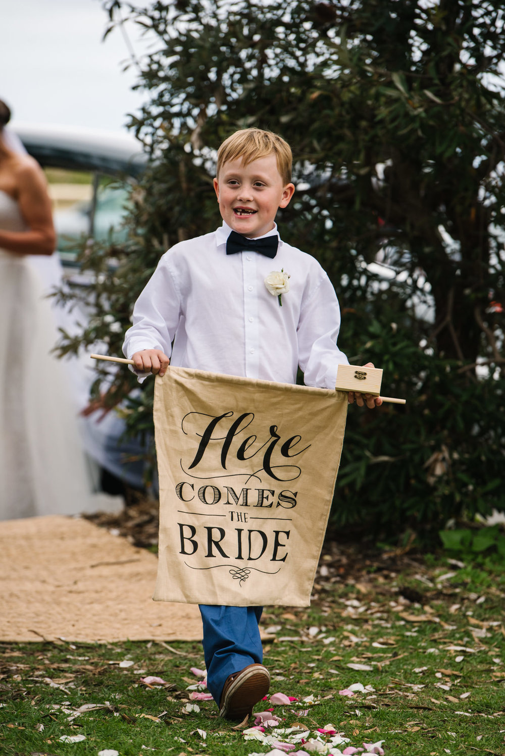 Ring bearer walking down aisle holding Here Comes the Bride sign