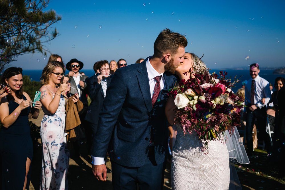 Bride and groom kiss as they exit their ceremony to guests blowing bubbles