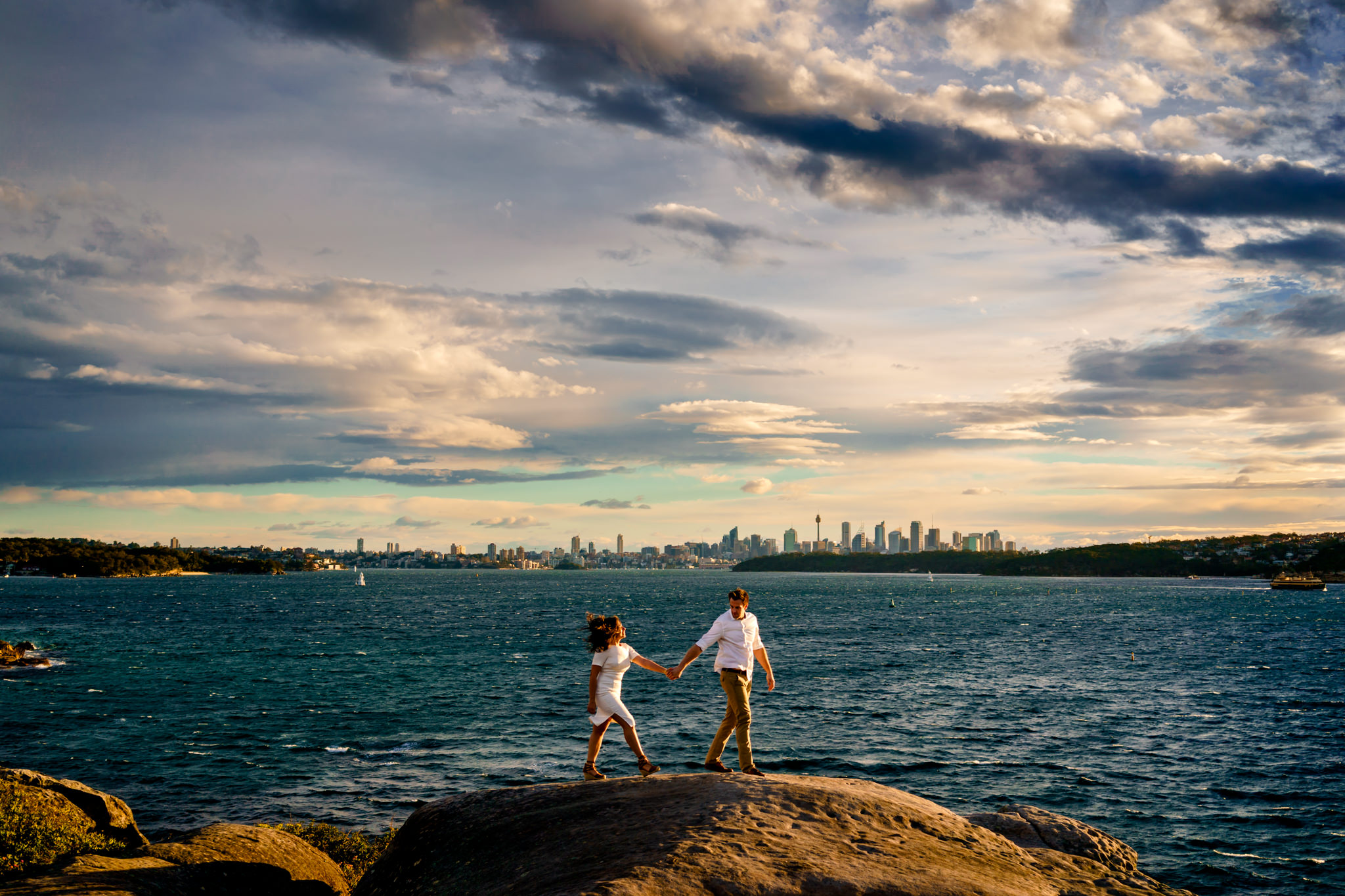 Engaged couple walk along rock cliffs with Sydney city and the harbour in the background in Watsons Bay NSW