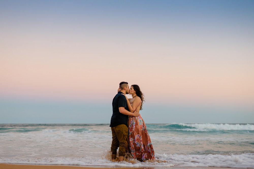 Engaged couple kissing in with their feet in the ocean and sunset colours in the sky behind them