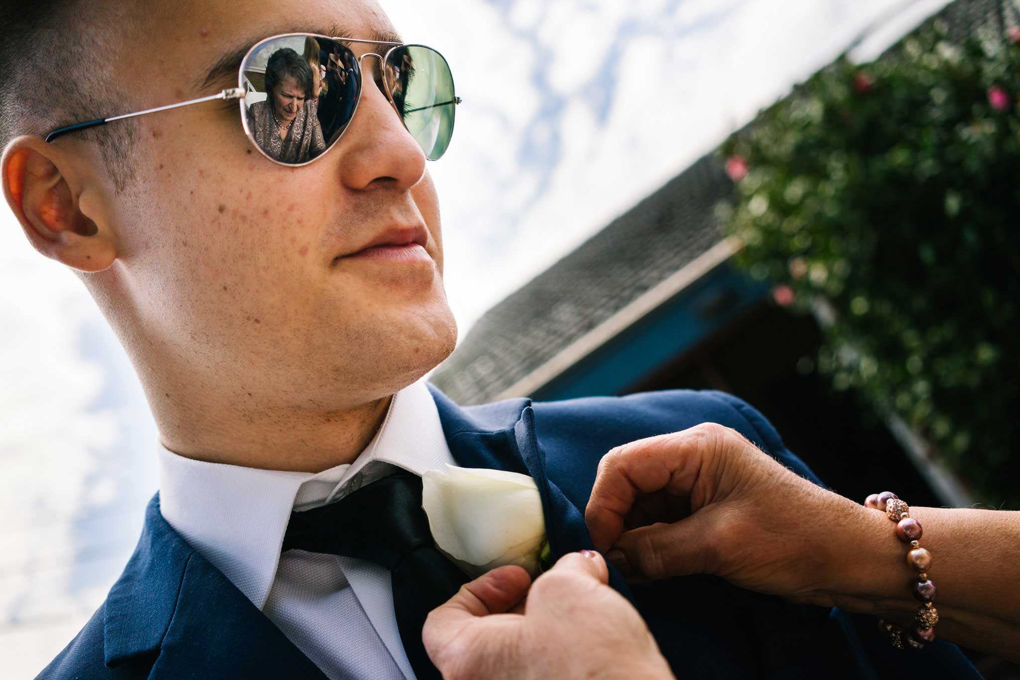 Reflection of grandmother in groomsman's sunglasses as she pins on buttonhole